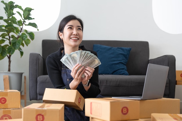 Happy woman freelancer working and holding money with cardboard box at home SME business online and delivery concept