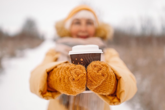 Happy woman hands in yellow knitted mittens holding hot chocolate or coffee for taking away on urban winter street