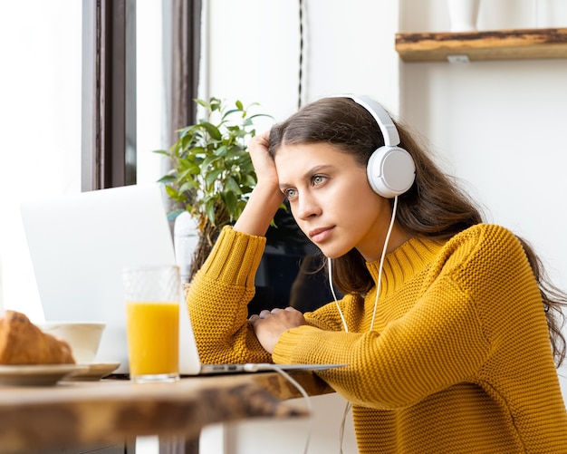 Happy woman in headphones studying online, using laptop and taking breakfast