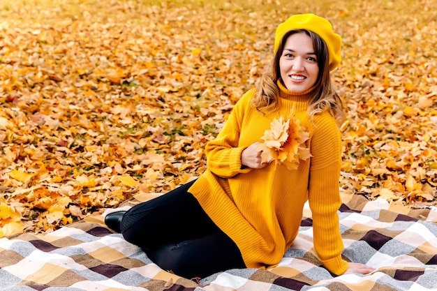 A happy woman hiding her eyes with a yellowed leaf in a yellow knitted beret with autumn leaves