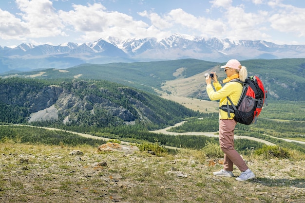 Happy woman hiker standing on the slope of mountain ridge against mountains blue cloudy sky on background Hiking woman taking pictures with digital camera