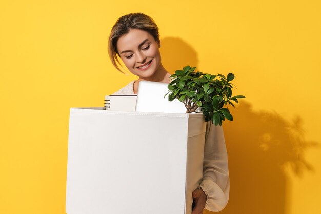 Happy woman holding box with personal items after job promotion against yellow background