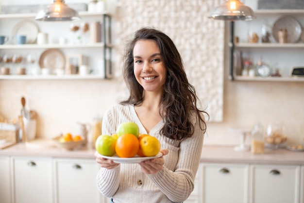 Happy woman holding plate with fresh fruits looking and smiling at camera standing in kitchen