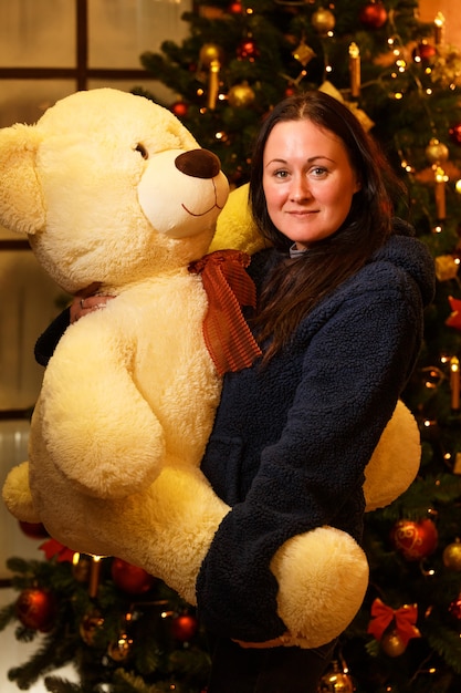 Happy woman holds a large stuffed bear toy in her arms near Christmas tree