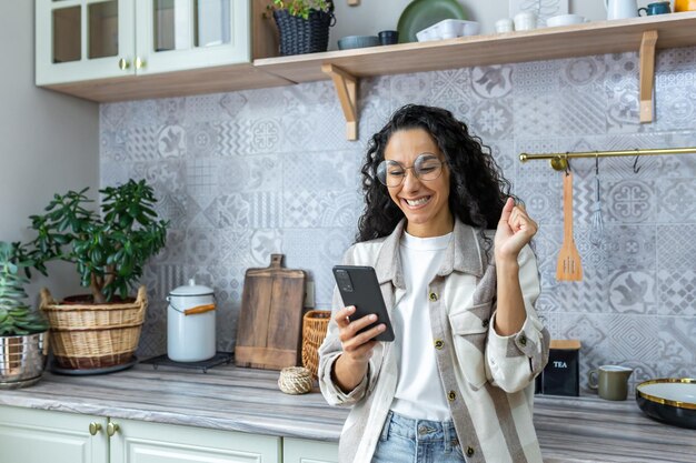 Happy woman at home in kitchen reading news online on phone hispanic woman using smartphone