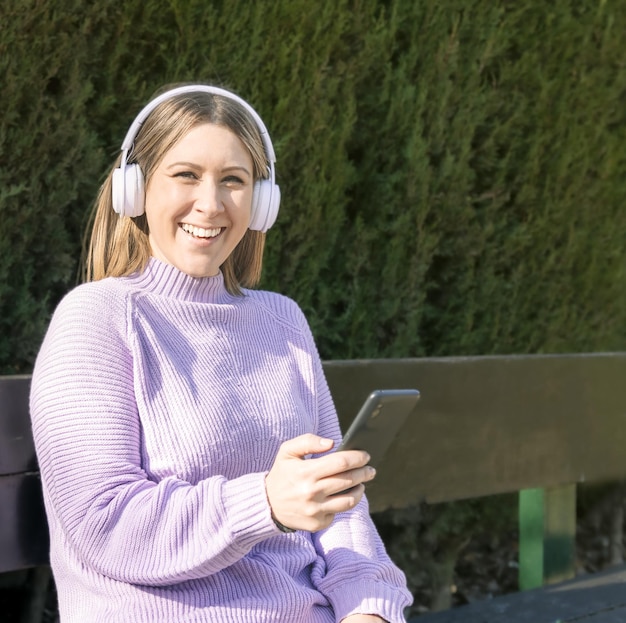 Happy woman is listening to music with wireless headphones sitting on a park bench using her smartphone