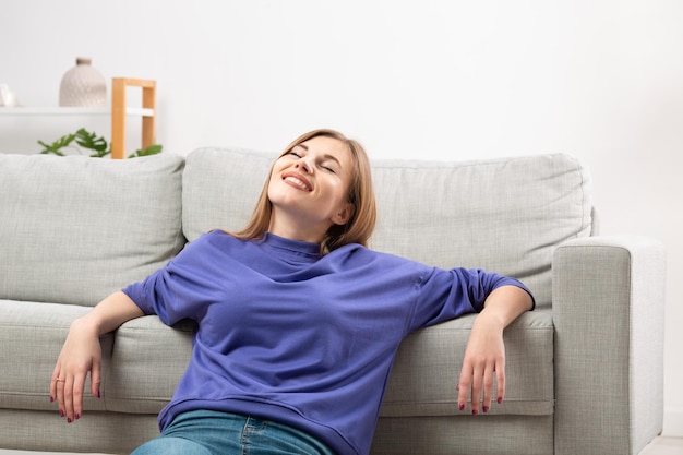 A happy woman is relaxing on the sofa breathing cool air from the air conditioner enjoying the comfortable climate in the ventilated living room meditating with her eyes closed Home comfort