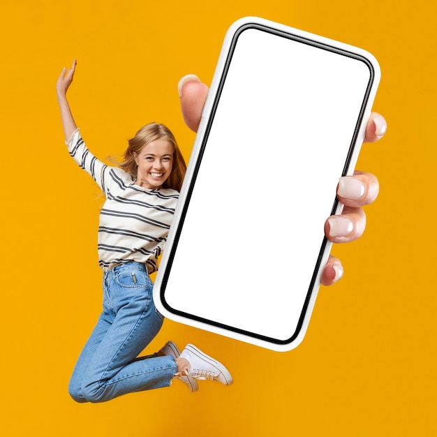 Happy woman jumping in air on orange background showing smartphone