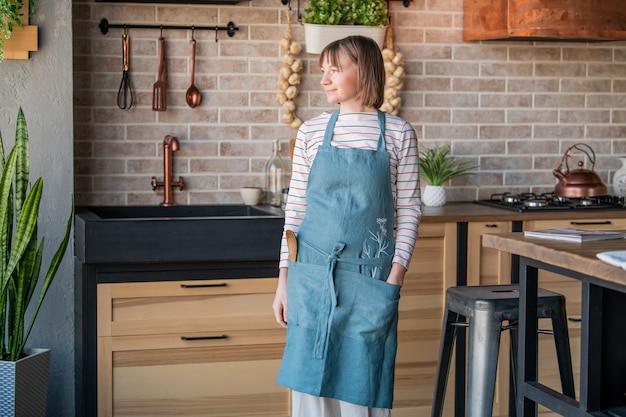 Happy woman in a linen apron standing in the kitchen at the table with a smile looks out the window