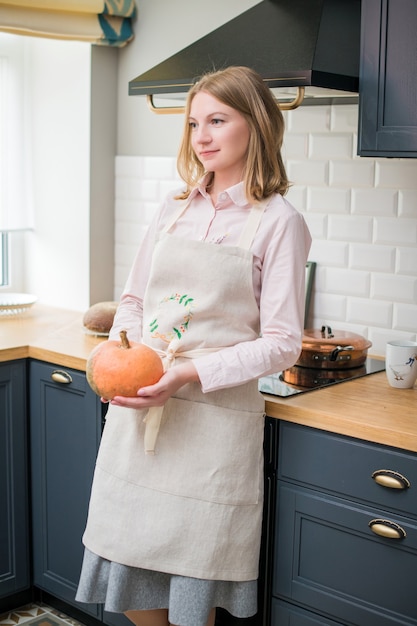 Happy woman in a linen apron stands in the kitchen next to the stove and holds a pumpkin in her hands