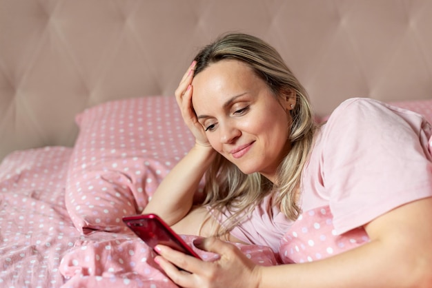 Happy woman lying in bed with smart phone in hands Waking up going to bed Sleep issues Young woman using smart phone text mesaging or take selfie on the bed at home