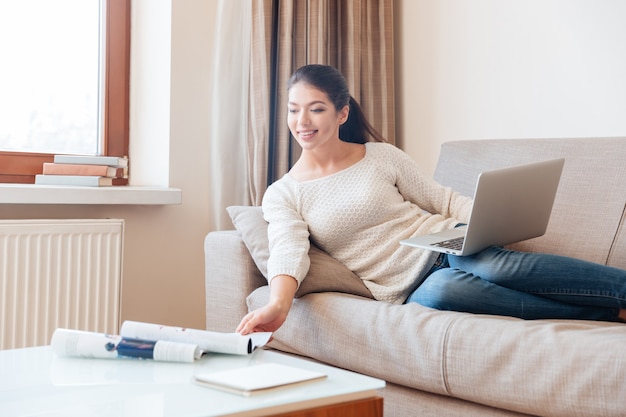 Happy woman lying on the sofa with laptop computer and stretching hand to take magazine