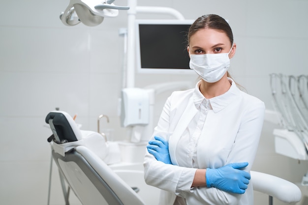 Happy woman in madical uniform and sterile gloves and mask is waiting for patients in modern office.