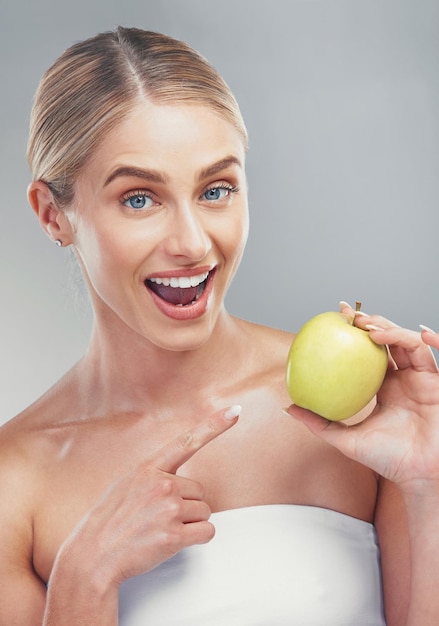 Happy woman pointing to green apple on studio background for healthy skincare vegan diet and lifestyle nutrition Portrait of smile female model holding fruit for wellness detox snack and vitamins