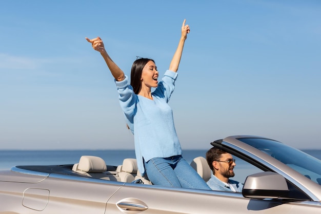 Happy woman raising hands riding in convertible with man