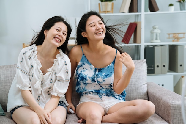 happy woman relaxing on sofa with air cooler machine breeze wind blowing hair. cheerful two lady best friends smiling enjoy electric fan turning on in living room hot indoors. summer heat concept.