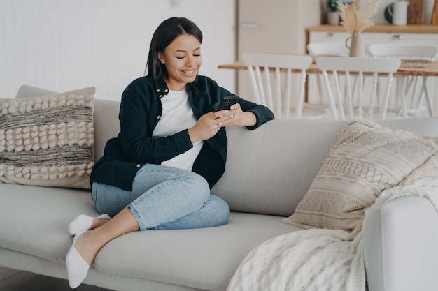 Happy woman relaxing with mobile phone and reading messages on comfortable couch in living room