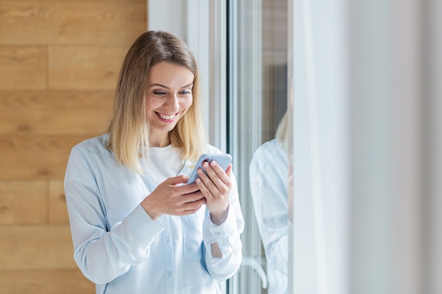 happy woman saw the good news on smartphone standing by the window in office or at home indoors