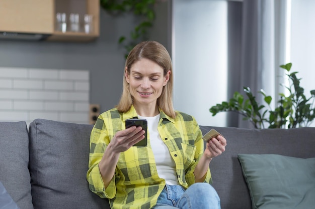 Happy woman shopping online sitting at home on the couch using phone and bank credit card