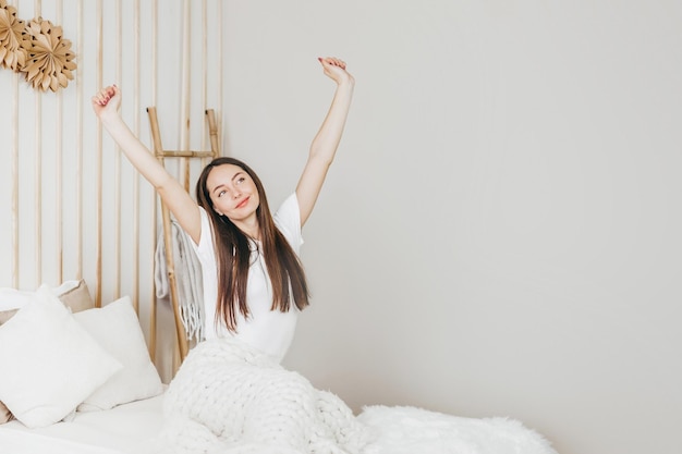 Happy woman sitting on the bed stretching after sleep and smiling in her room