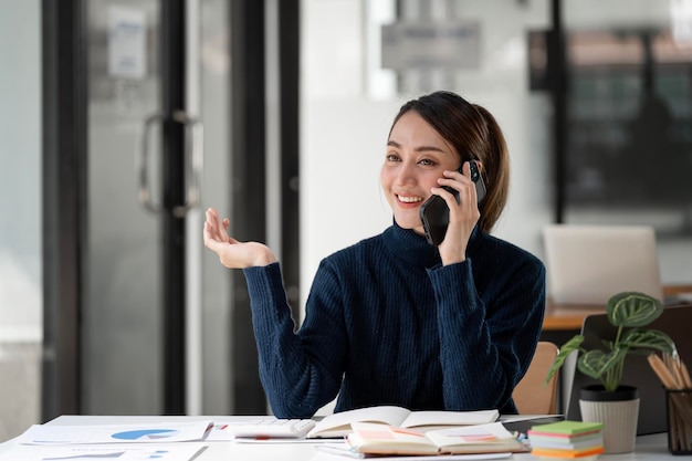 Happy woman sitting at desk behind her laptop and talking with somebody on her mobile phone while working at open office