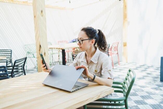 Happy woman sitting at outdoor cafe table and talking on phone with cup of coffee smiling woman enjoying telecommuting in cafe or studying online