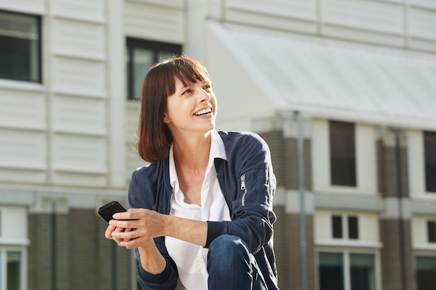 Happy woman sitting outside in city with smart phone