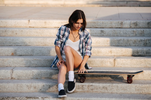 Happy woman sitting on the stairs looking down at her skateboard with care