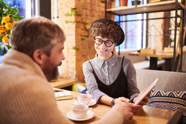 Happy woman in smart casual and her boyfriend watching funny video in touchpad while relaxing in cafe