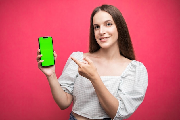 Happy woman standing and hold smartphone and point at phone on pink background. Green screen phone s