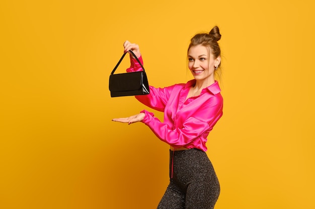 Happy woman stands in profile and holds her purse at arm against yellow studio background