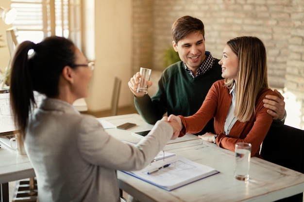 Happy woman talking to her husband while shaking hands with their financial advisor in the office