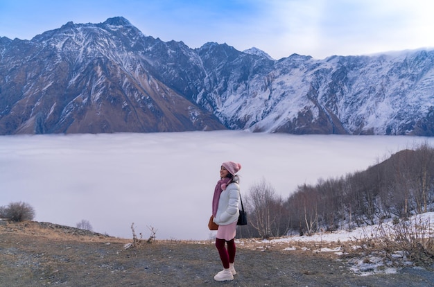 Happy woman tourist with fantastic scenery of the Caucasus mountains and sea of mist in winter near Gergeti Trinity Church in Kazbegi Georgia