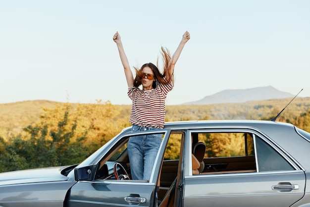 Happy woman traveler climbed on the car and spread her arms smiling happily looks at the nature around Lifestyle in travel and joy