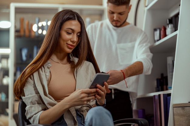 Happy woman using smart phone while getting her hair styled at beauty salon