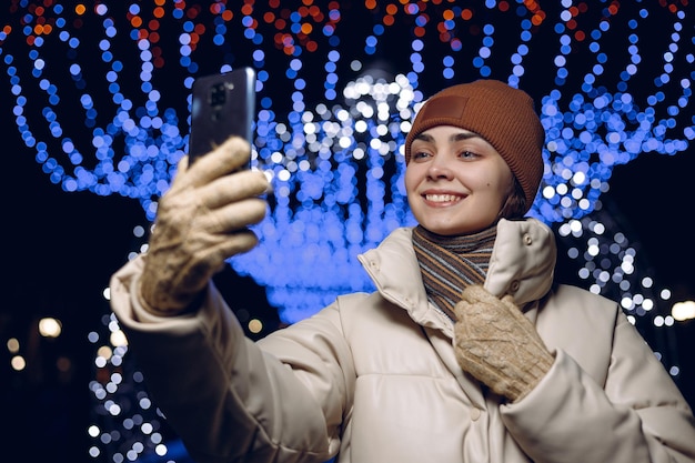 Happy woman in warm clothes taking selfie on mobile phone while standing in city