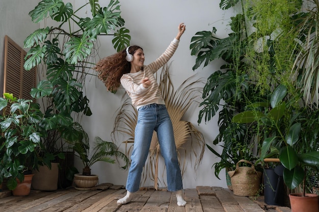 Happy woman wear wireless headphones listening to music dancing in home garden with tropical plants