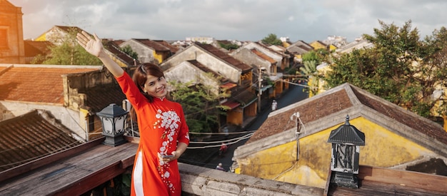 Happy woman wearing Ao Dai Vietnamese dress traveler sightseeing view at rooftop at Hoi An ancient town in Vietnam landmark and popular for tourist attractions Vietnam and Southeast travel concept