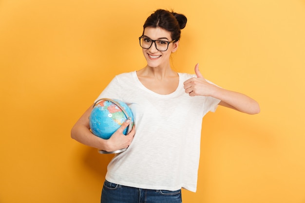 Happy woman wearing glasses holding globe showing thumbs up gesture.