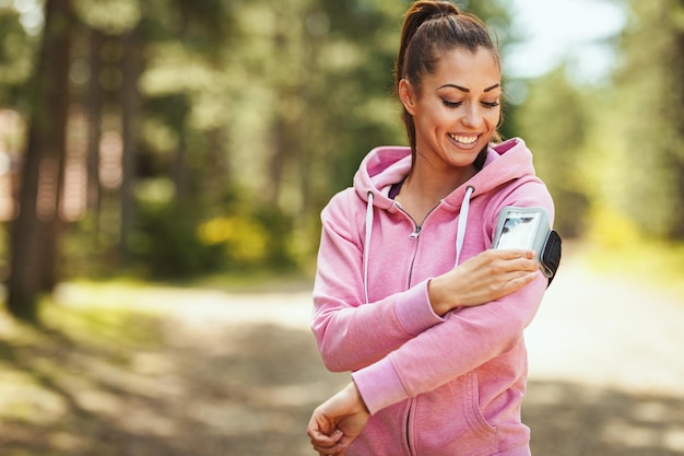 Happy woman, wearing sportswear, checking her smartphone in a break of exercises in a sunny forest, enjoying landscape among the trees.