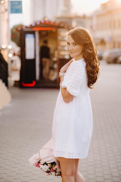 A happy woman in a white dress at sunset with a bouquet of flowers in the city