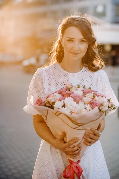 A happy woman in a white dress at sunset with a bouquet of flowers in the city