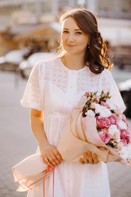 A happy woman in a white dress at sunset with a bouquet of flowers in the city