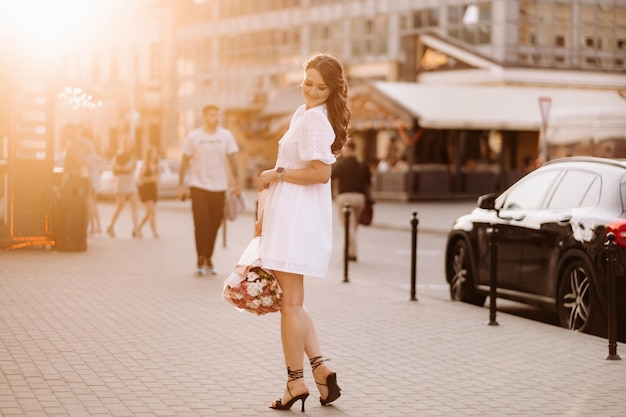 A happy woman in a white dress at sunset with a bouquet of flowers in the city