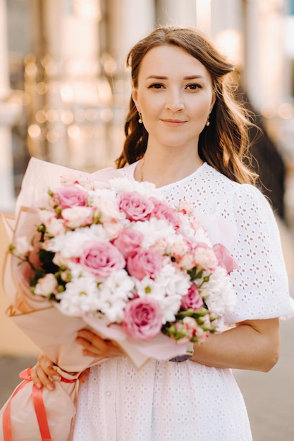 A happy woman in a white dress at sunset with a bouquet of flowers in the city