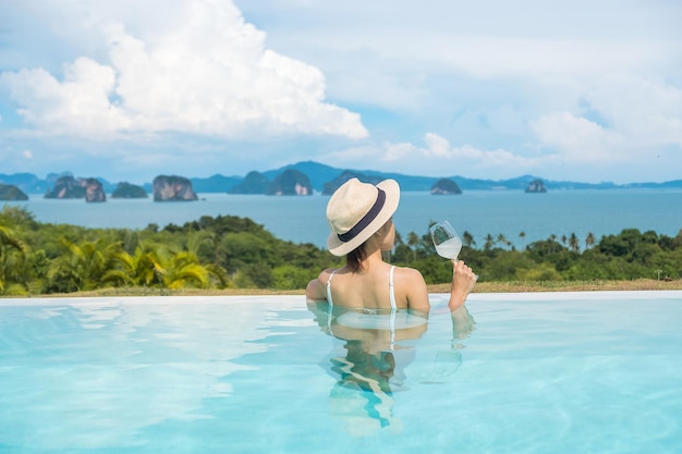 Happy woman in white swimsuit drinking wine in luxury swimming pool hotel against beautiful view young female with hat enjoy in tropical resort Relaxing summer travel holiday and vacation concept