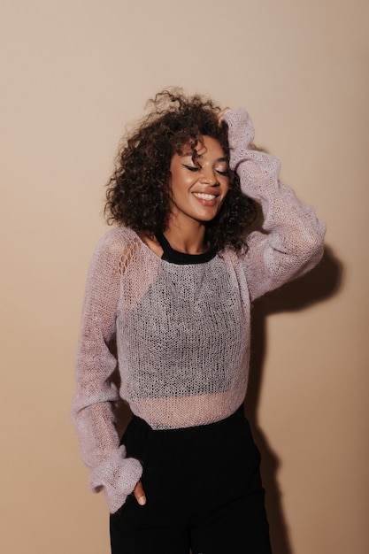 Happy woman with beautiful makeup and wavy dark hair in black top, pink sweater and dark pants smiling with closed eyes on beige wall..