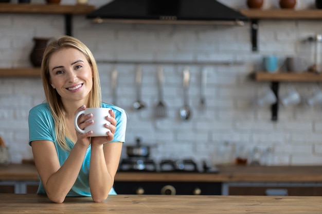 Photo happy woman with a cup of coffee looking at camera while standing in the kitchen at home