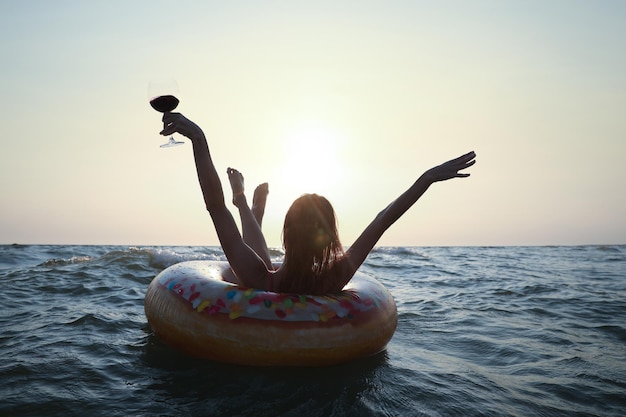 Happy woman with glass of wine and inflatable ring in sea at sunset back view