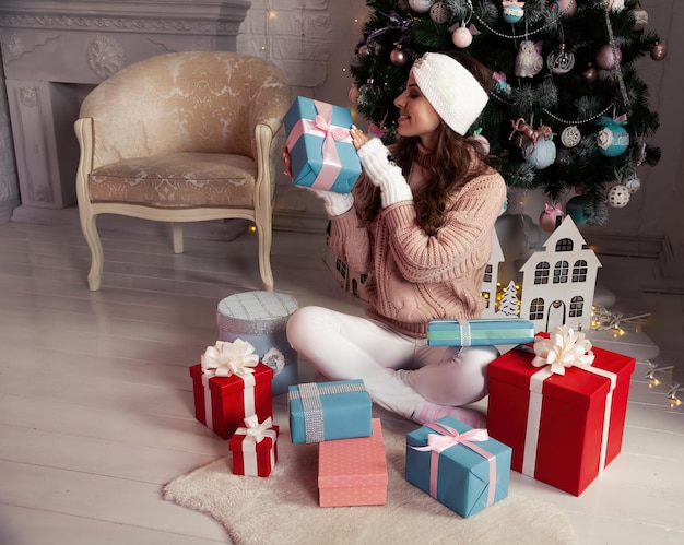 Happy woman with presents in front of the Christmas tree Stylish female in cozy knitted sweater holding present in festive room Merry Christmas and happy holidays New Year and Christmas concept